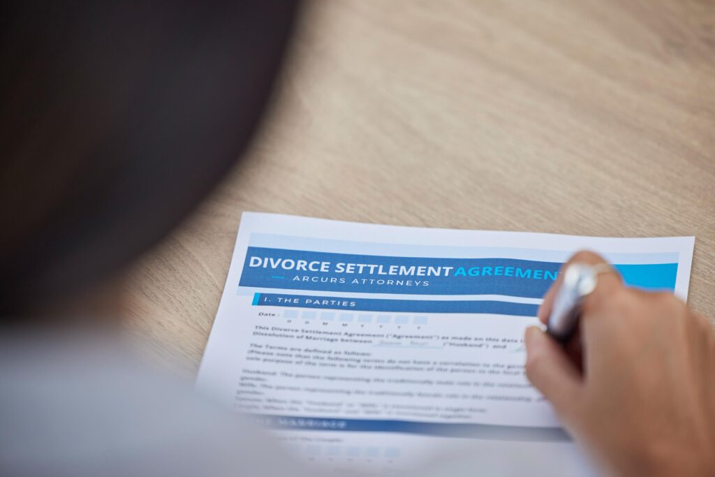 A hand holding a pen is shown in the action of signing a divorce settlement agreement on a wooden desk.