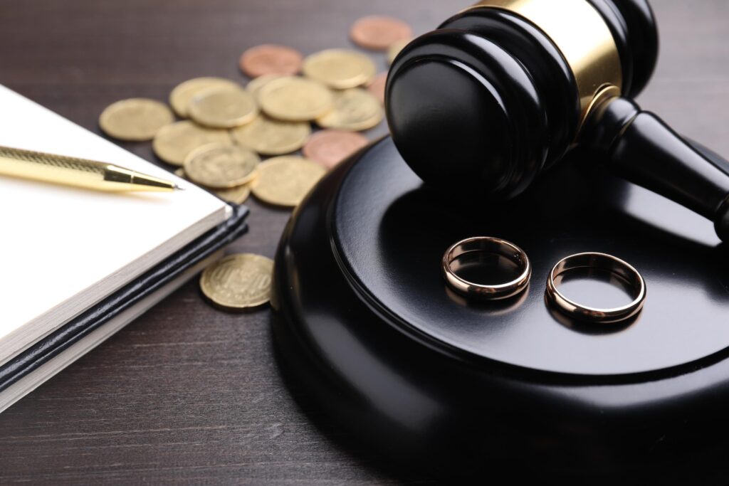 Alimony. Gavel, wedding rings, notebook and coins on wooden table, closeup
